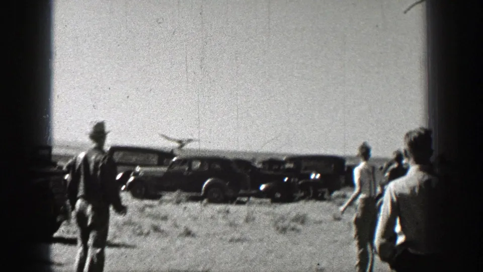 In 1938 Washington home-movie footage, men watch a small model airplane over a dusty field with dark period cars behind them.