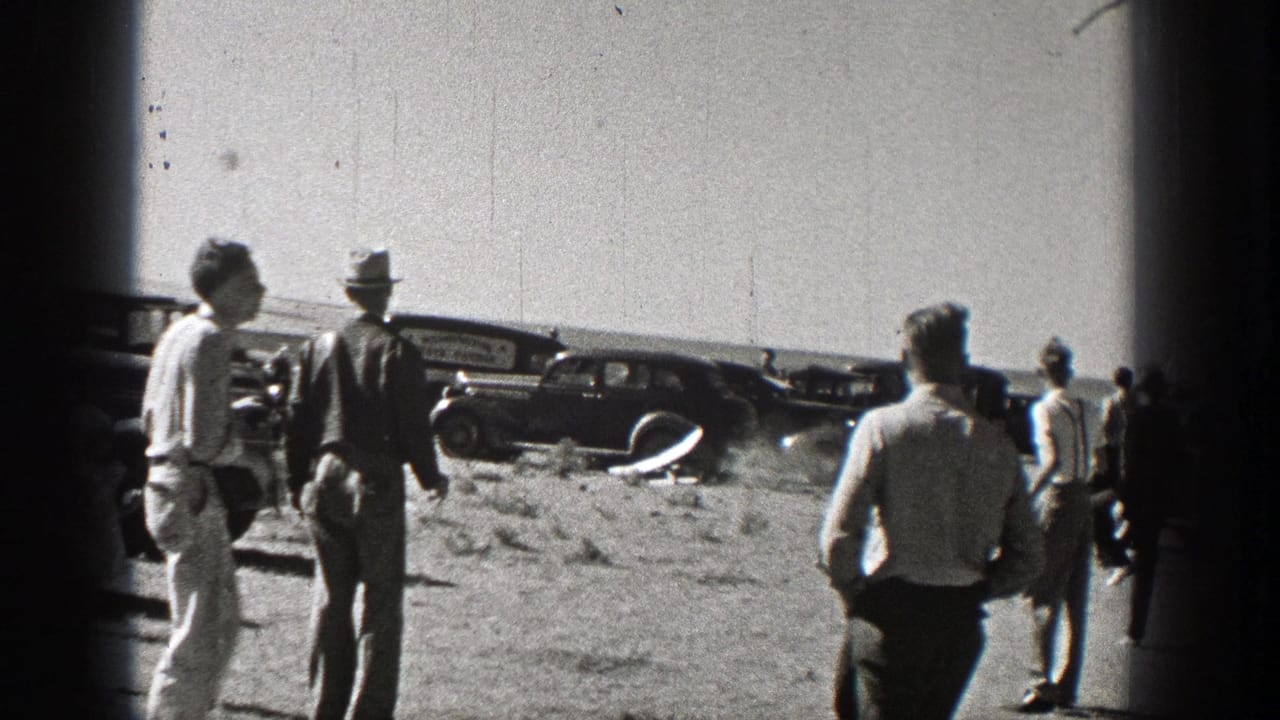Two men stand in the foreground while others look toward a small airplane near parked cars.