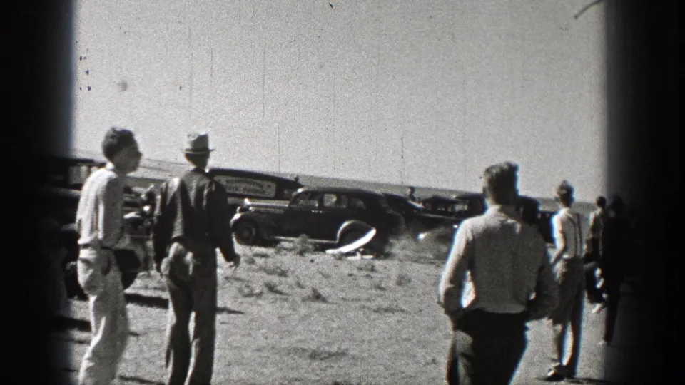 Men in 1930s attire stand in a Washington field watching a small model airplane near parked late-1930s cars.
