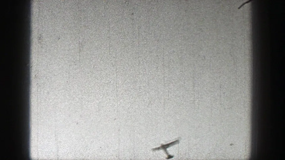 The model airplane is seen low in the frame against a washed, grainy field area in the 1938 home-movie sequence.