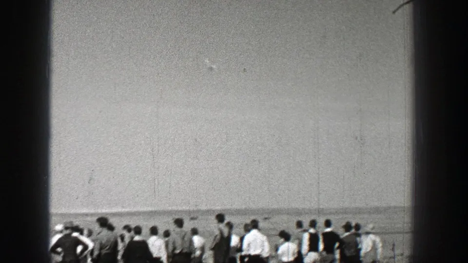 Washington 1938 frame with a low crowd line in an open field beneath a wide sky, watching aircraft activity off-frame or overhead.
