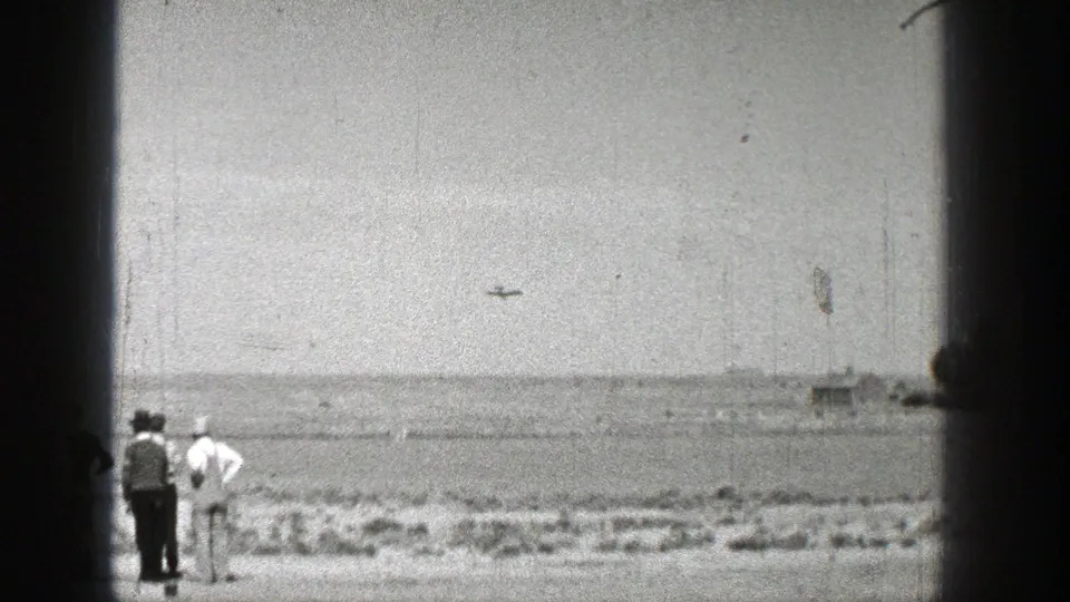 Washington 1938 home-movie frame showing two spectators in the foreground and a small aircraft crossing the distant sky.