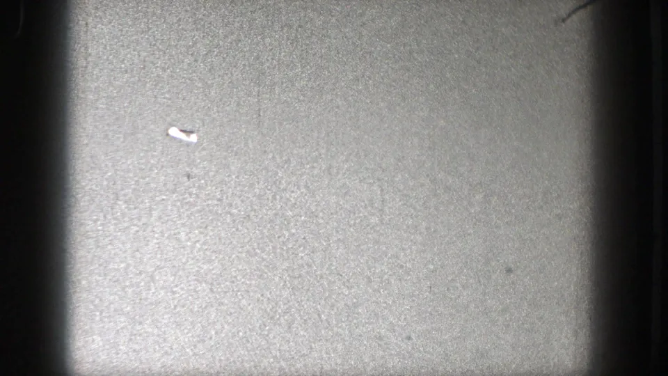 The aircraft remains distant in a soft, grainy sky frame, partially framed by dark vertical 8mm edge shadows.
