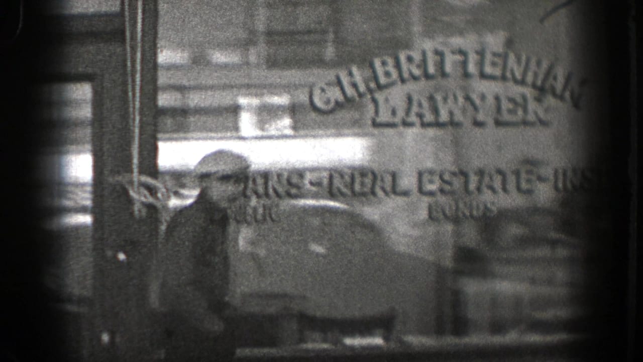 Man in a cap stands near a storefront window with painted lettering behind him.