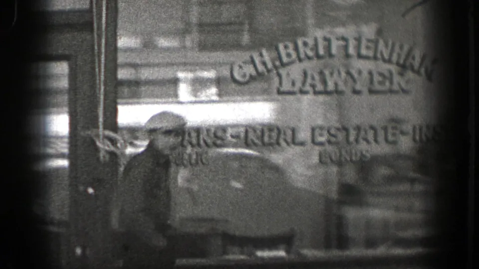 Black-and-white 8mm frame from Washington showing a man near a storefront window with partially readable real-estate lettering.