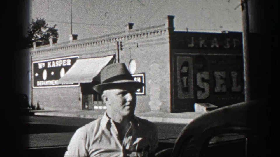 Washington 1938 home-movie frame showing a man in a fedora beside a car, with storefront signs and awnings across the street.