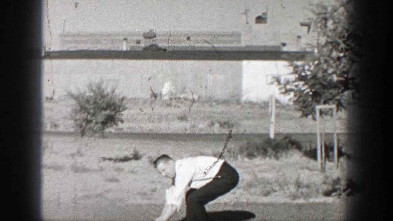 Man in a white shirt bends toward the ground in a grassy area beside a low building.
