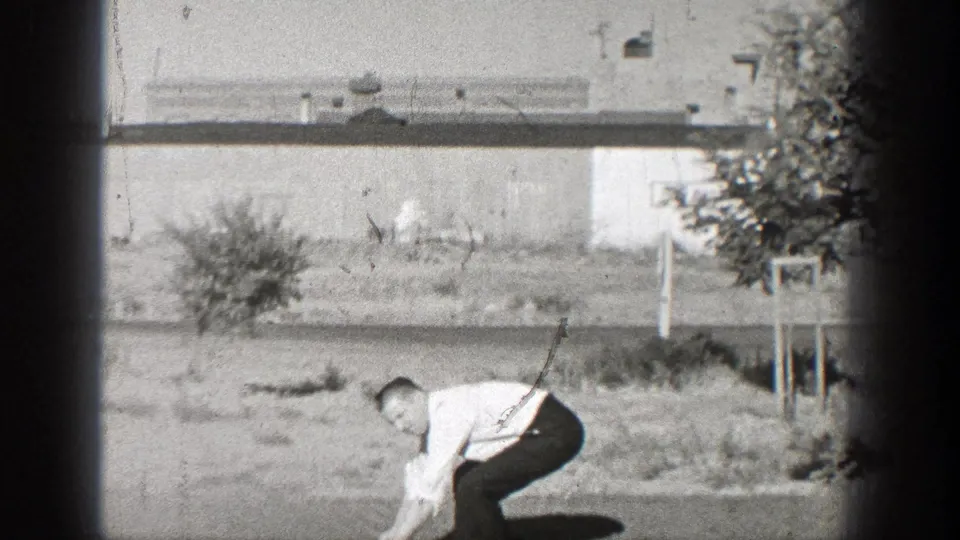 In 1938 Washington footage, a man in a white shirt and dark trousers bends down near grass, a small tree, and a low building.