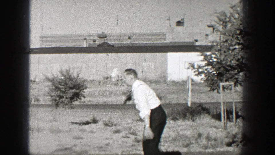 A man in a white shirt and dark trousers moves across a grassy Washington lot with a low building and young tree in the background.