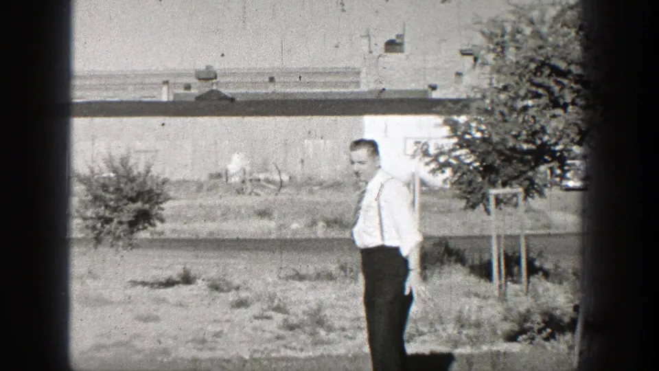 A man in a white shirt and dark trousers stands in a grassy area in Washington, with a low building, tree, and open ground behind him.