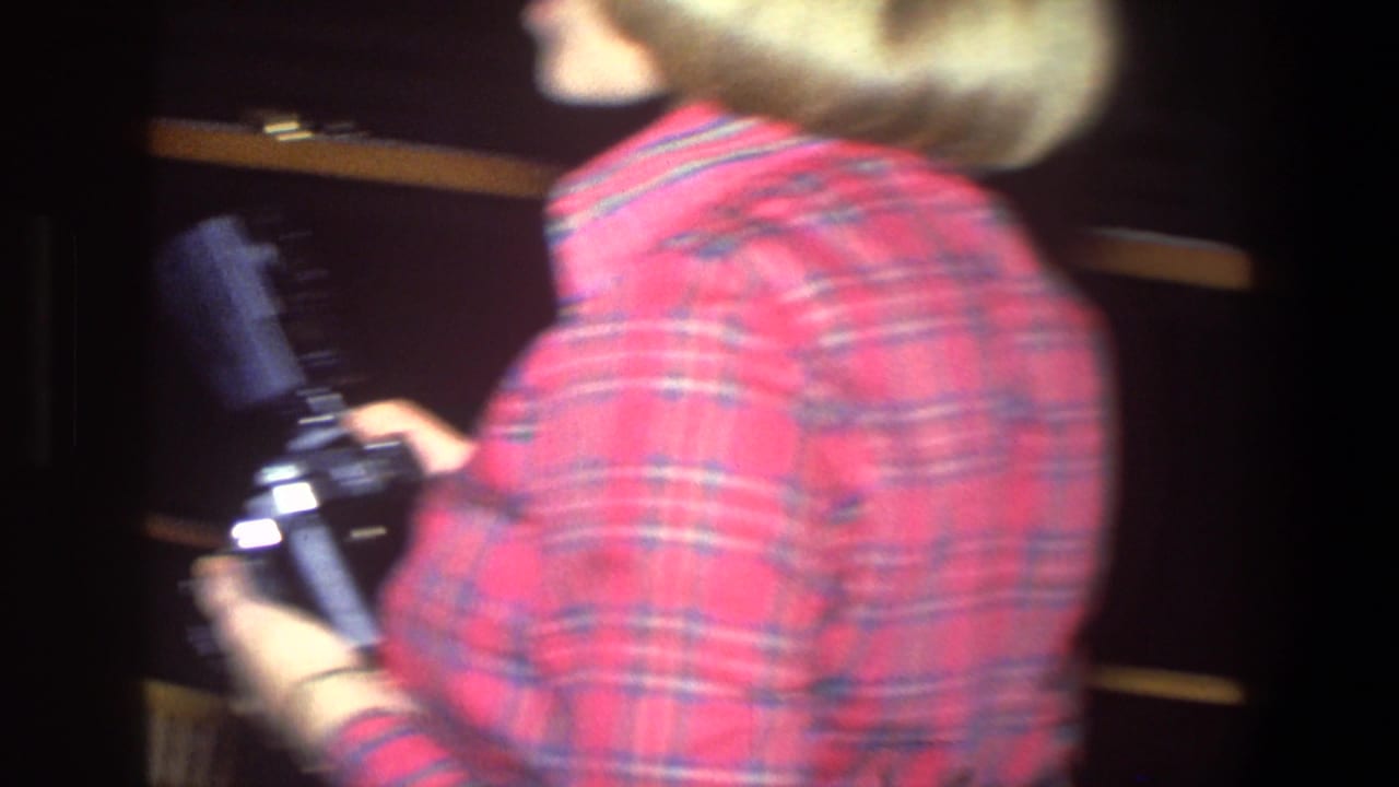 Woman in a red plaid shirt turns while holding a camera in a dim interior.
