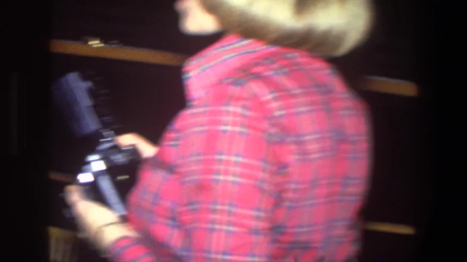 In a 1971 Saint Paul home-movie frame, a woman in a red plaid shirt turns with a camera near dark shelves and wood trim.