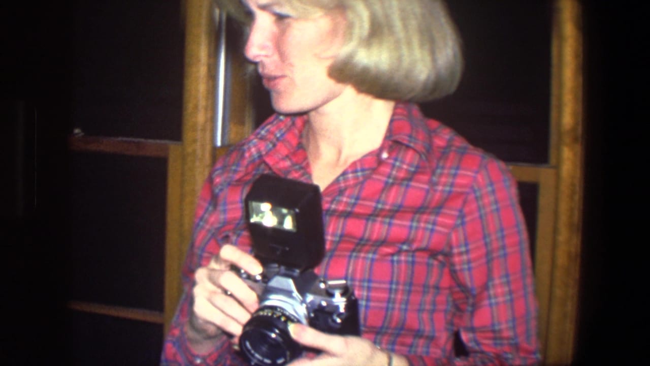 Woman in a red plaid shirt holds a still camera with a flash attachment.