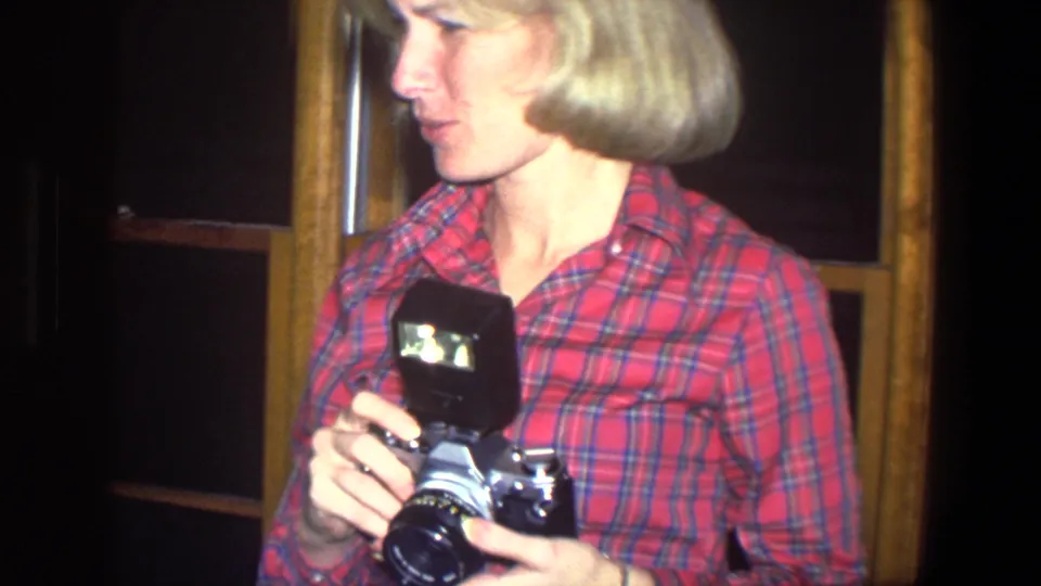 A woman in a red plaid shirt holds a vintage still camera with a flash unit inside a Saint Paul home in 1971.