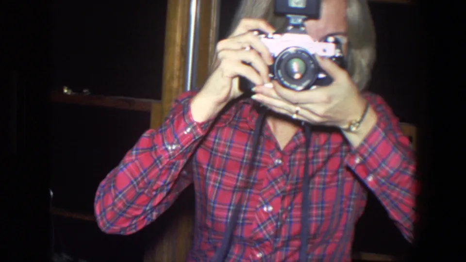 The woman lifts a vintage camera to eye level, with the flash unit visible above the camera body in a 1971 home interior.