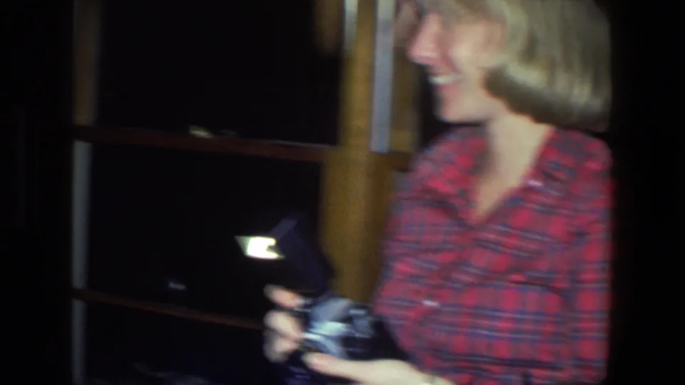 A smiling woman in a red plaid shirt holds a camera and flash unit in a softly focused 1971 Saint Paul home-movie frame.