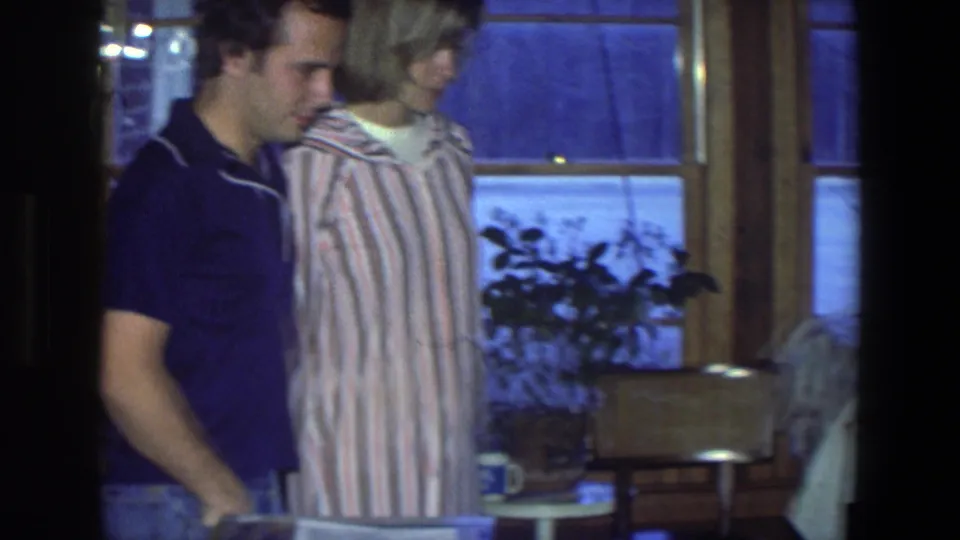 The couple looks down out of frame in a 1971 Saint Paul interior, with snow, plants and a small table visible.