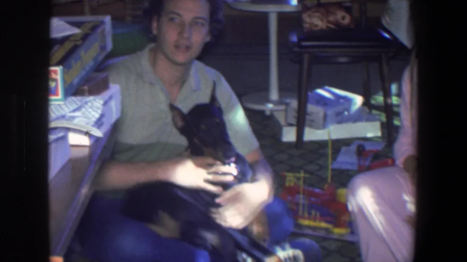 A young man in a green shirt holds the puppy on a Saint Paul living-room floor, with toys and household items visible around him.