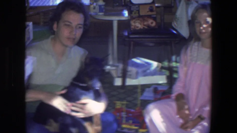 The home-movie frame shows the man with the puppy on the floor while a seated woman in light clothing appears beside toys in the room.