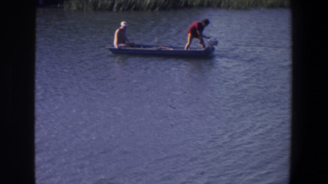 Two men in a small boat on rippled lake water, one seated at the bow and one standing near the motor.