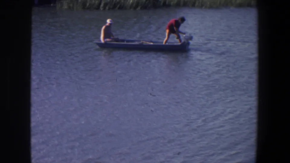 1971 Saint Paul home-movie frame showing two adult men in a small boat on a lake, with one seated forward and one bent near the stern motor.