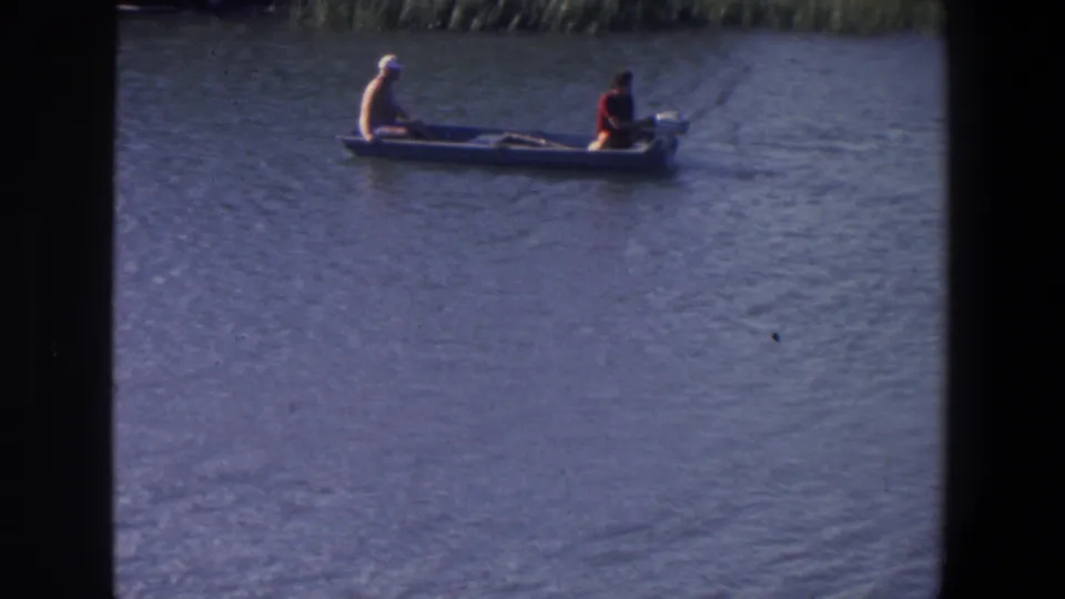 1971 Saint Paul frame showing two adult men in a small boat on a lake, with shoreline reeds faintly visible in the background.