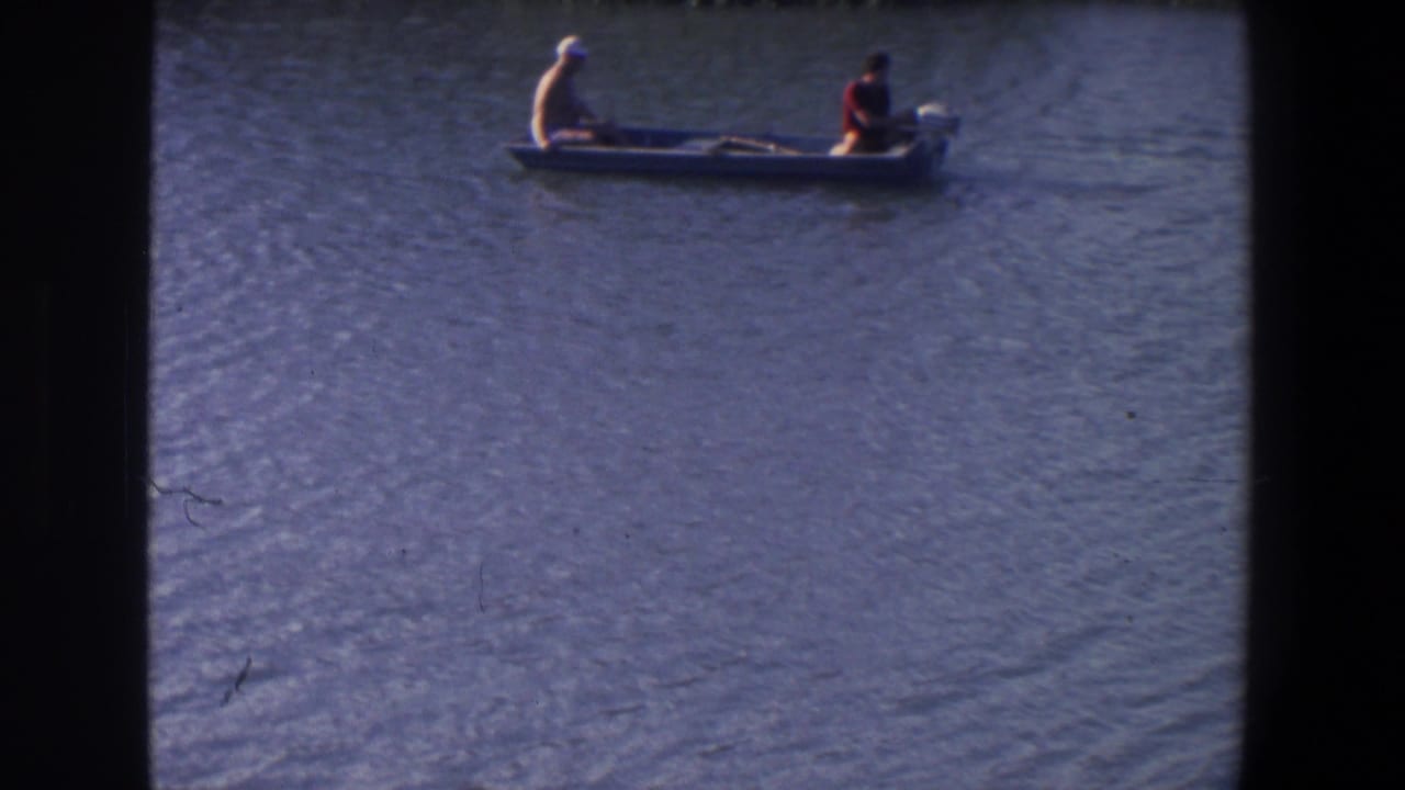 Two people in a small motorboat move across rippled water near a dark shoreline.