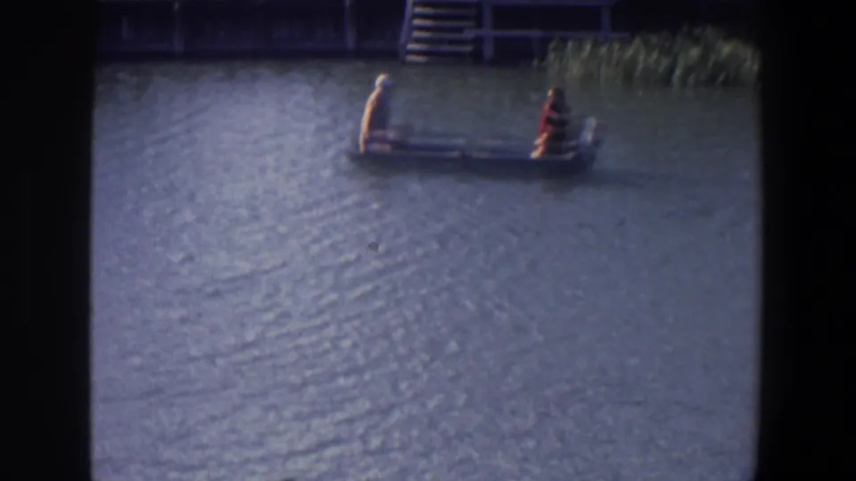 A distant 1971 frame shows two people seated in a small motorboat near waterside steps and reeds in Saint Paul.