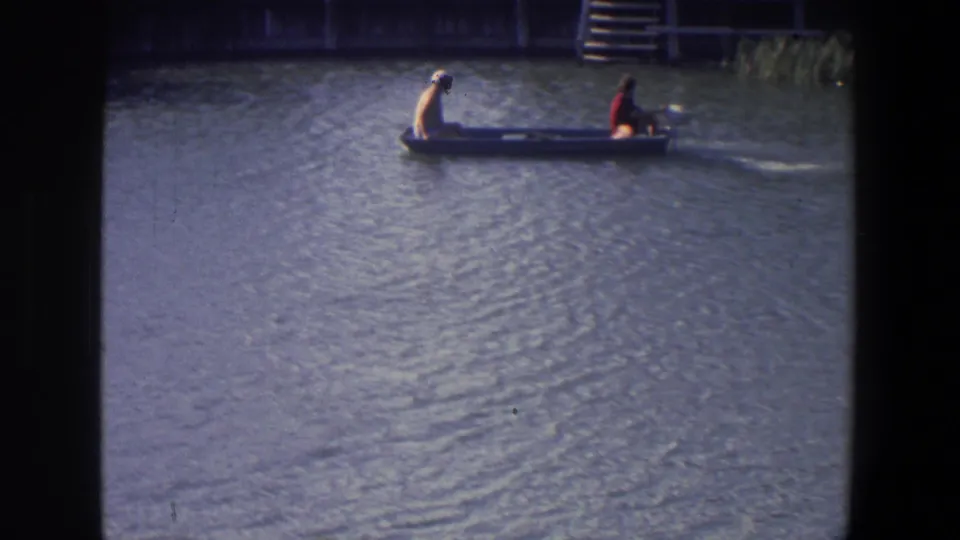 In 1971 Saint Paul footage, two people sit in a small motorboat moving across calm lake water with dock structures in the background.