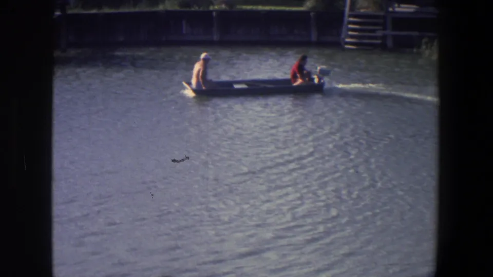 The small boat continues across the Saint Paul lake scene, with one person near the bow and another operating the motor at the stern.