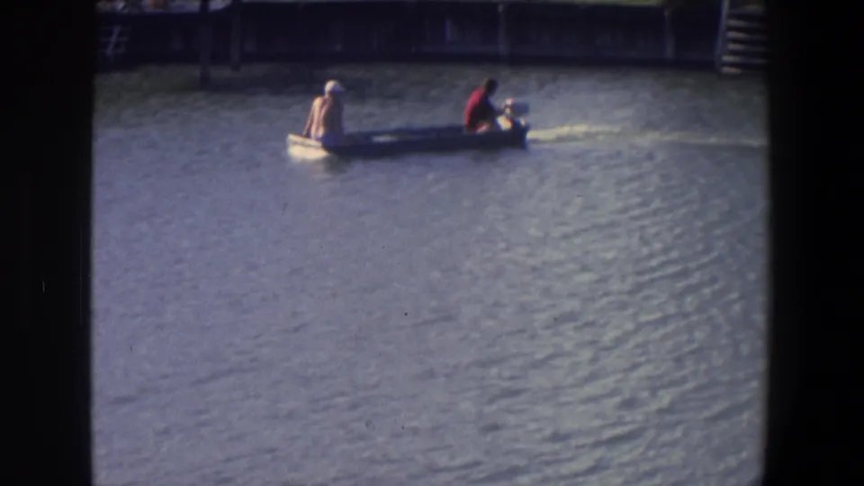The motorboat passes closer across the frame in 1971 Saint Paul home-movie footage, with rippled water and dock steps behind it.