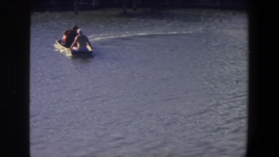 The boat angles toward the camera in the Saint Paul lake scene, showing two seated people, a trailing wake, and broad rippled water.