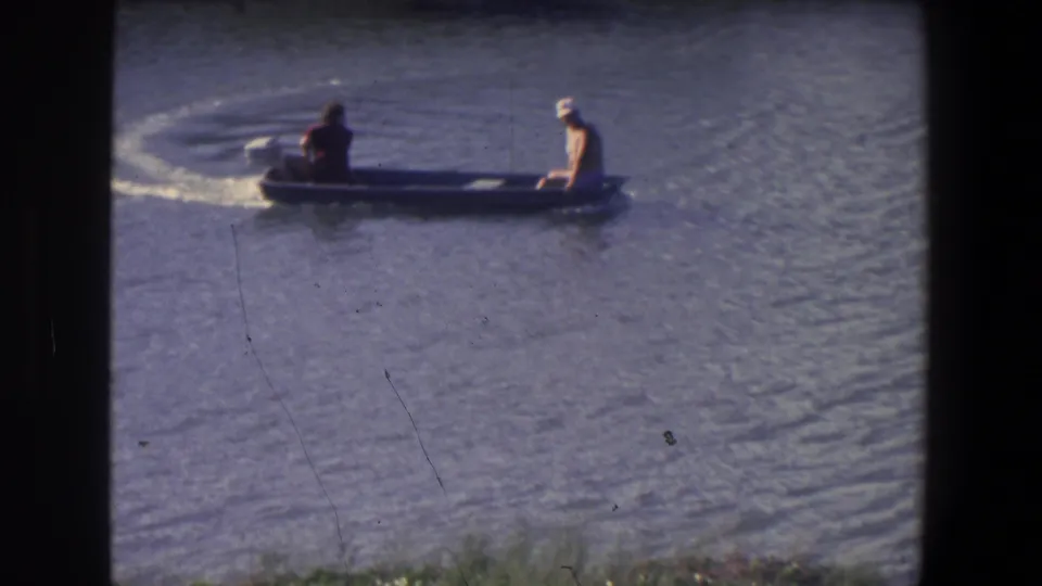 Two people in a low motorboat pass through the frame on rippled water in 1971 home-movie footage.