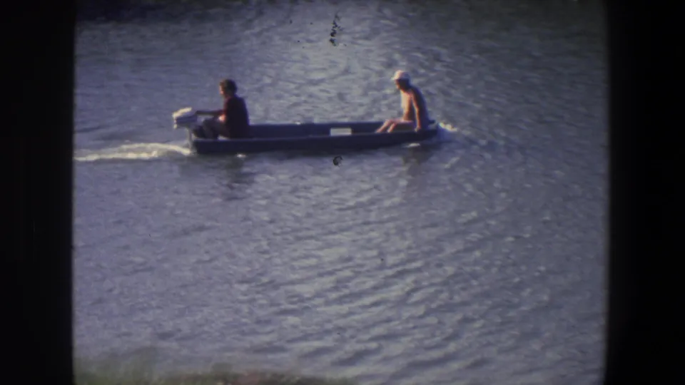 In 1971 Saint Paul footage, two men travel across calm water in a small gray motorboat with an outboard motor.