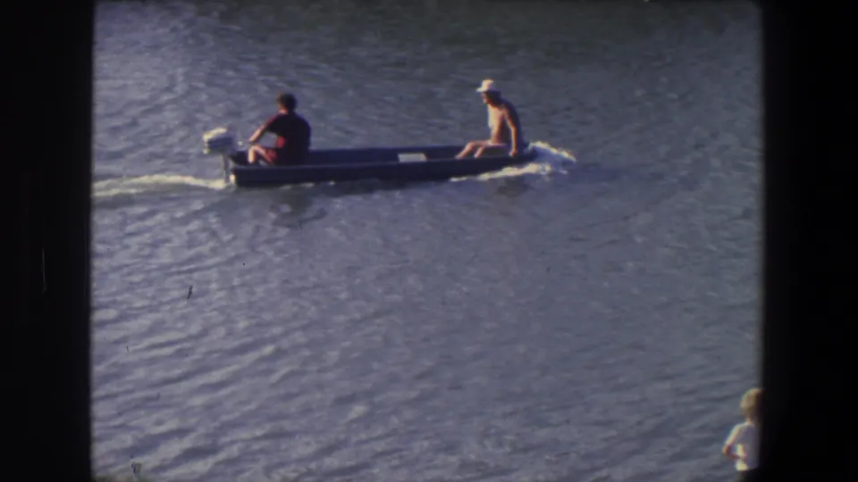 The gray motorboat continues across sunlit water, with one man at the motor and another seated near the stern in a white cap.