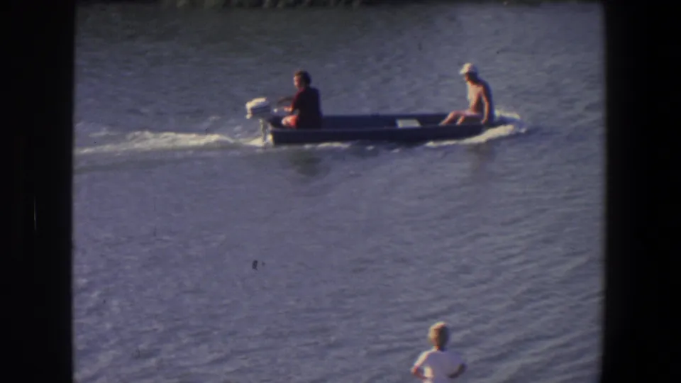 A 1971 home-movie frame shows the two men and gray outboard motorboat moving across open water with soft 8mm grain.