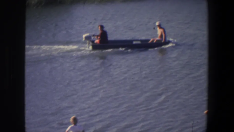 The boat carries two men across rippled water, with a distant shoreline figure partly visible near the lower edge of the frame.