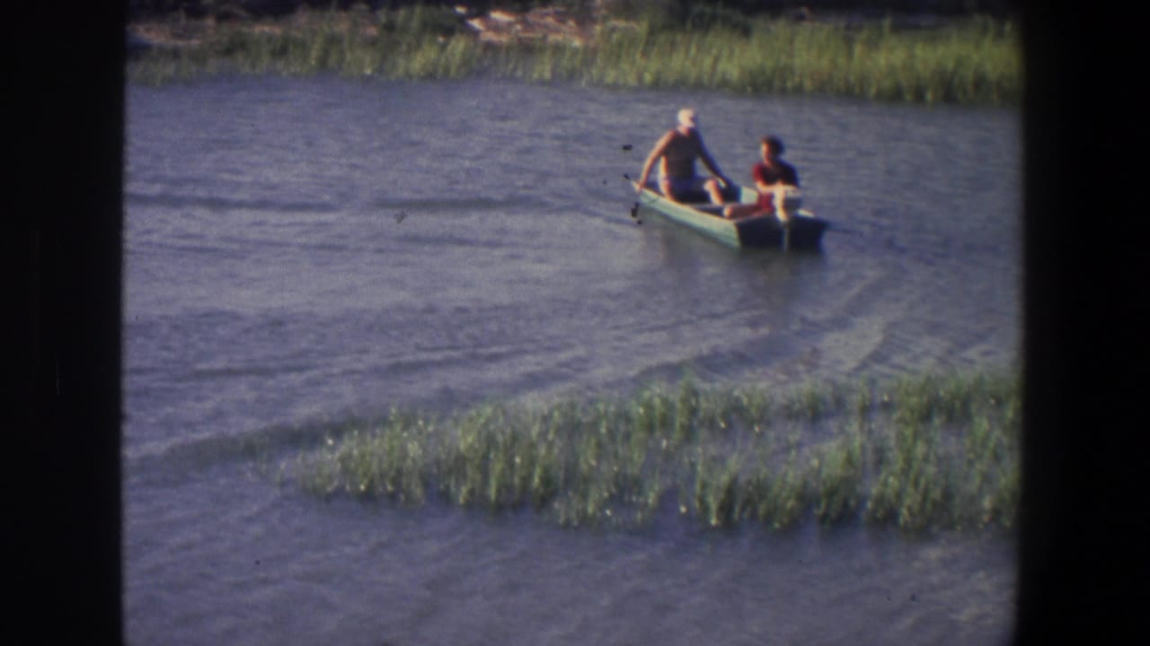Two people in a small boat move through rippled water near marsh grasses.