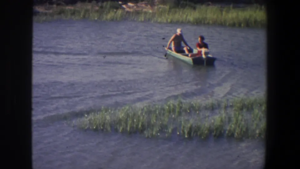 In 1971 Saint Paul home-movie footage, two people sit in a small boat on calm water bordered by reeds.