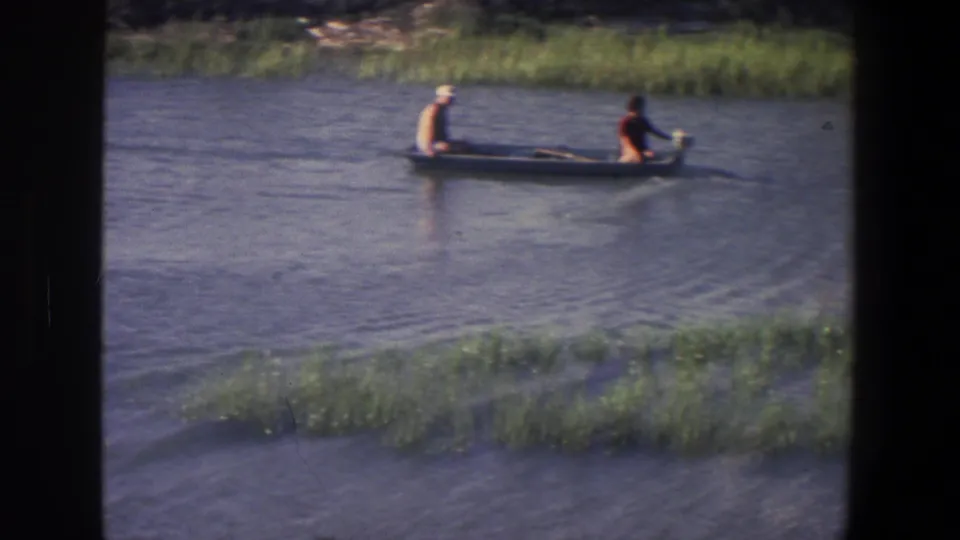 A man in a light cap and a person in a red top sit in a small boat on calm water in the Saint Paul, Minnesota clip.