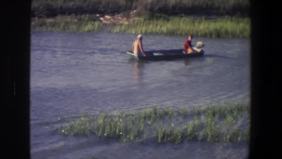 The small boat continues past marsh vegetation, with one person near the front and another near the motor area at the rear.