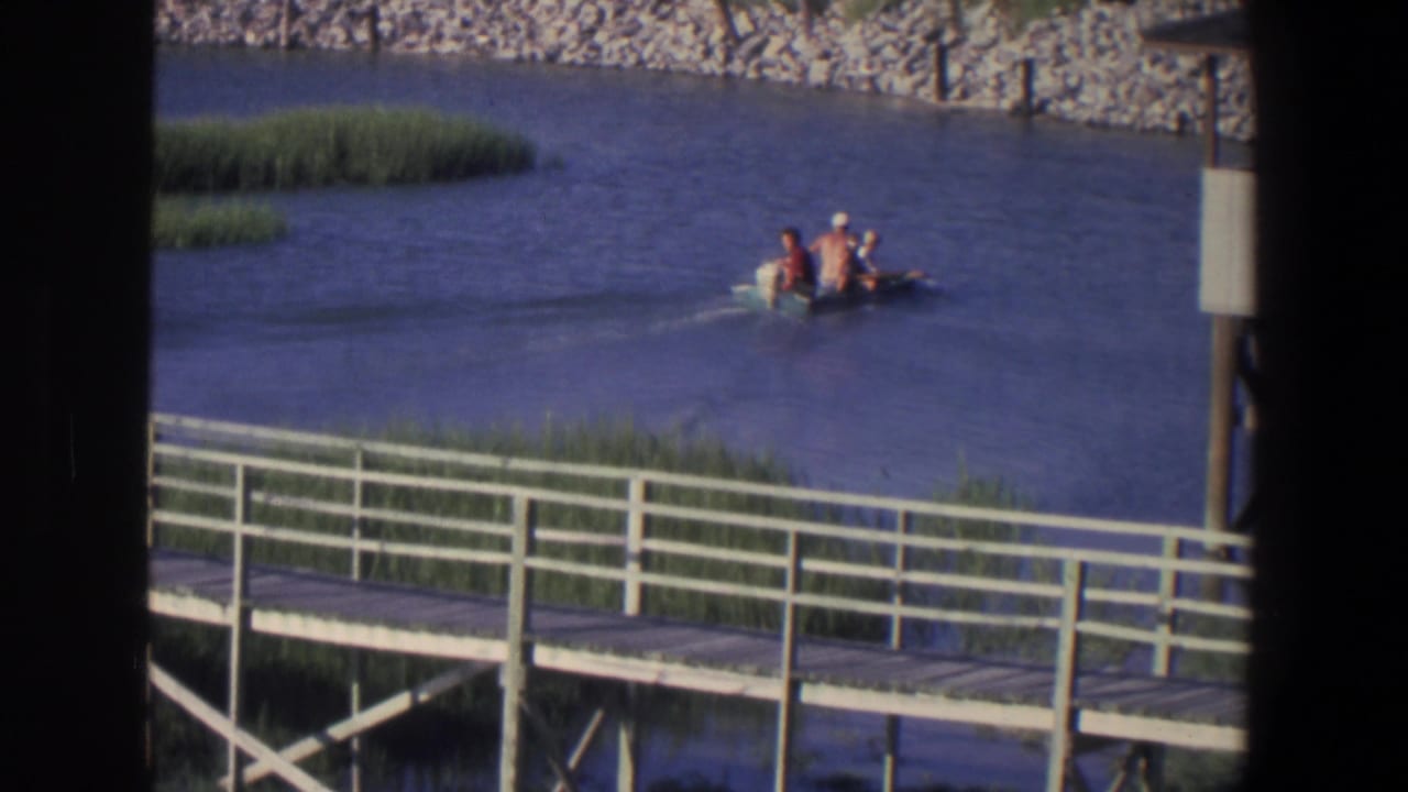 Small motorboat with three people on a calm river near a white-railed boardwalk.