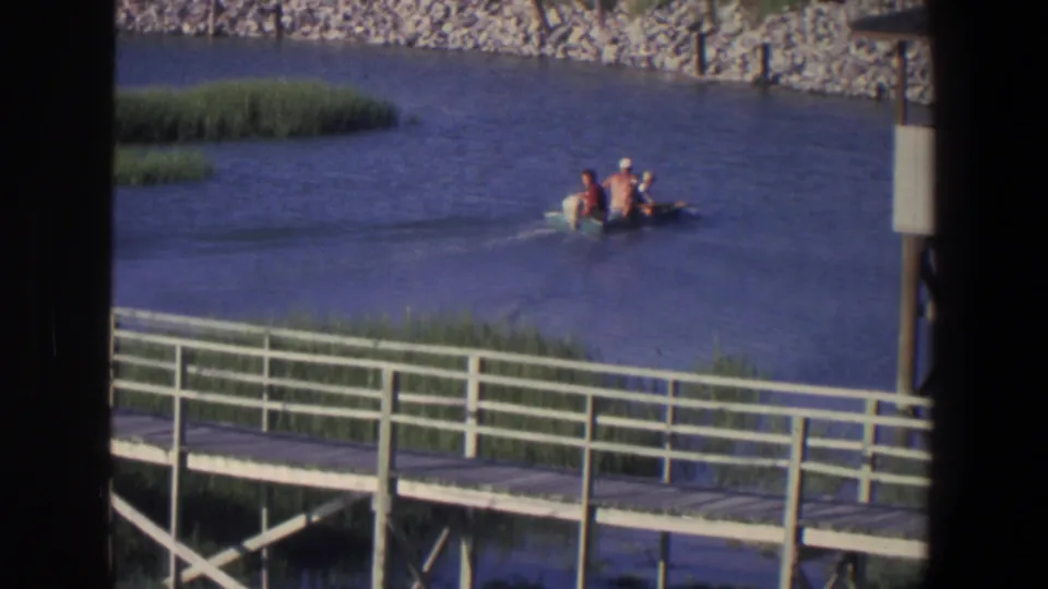 1971 Saint Paul home-movie frame showing three people in a small motorboat near reeds, a boardwalk, and a rocky riverbank.