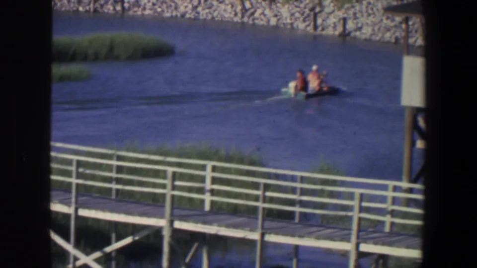 1971 Saint Paul river scene with a small motorboat carrying three people past reeds and a white-railed walkway.