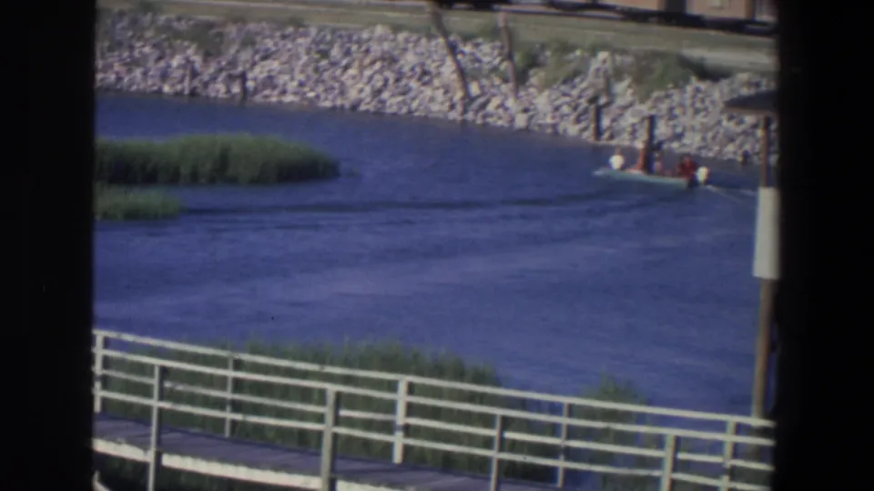 1971 Saint Paul home-movie frame with the motorboat partly at frame edge, reeds, river water, and a white boardwalk in front.