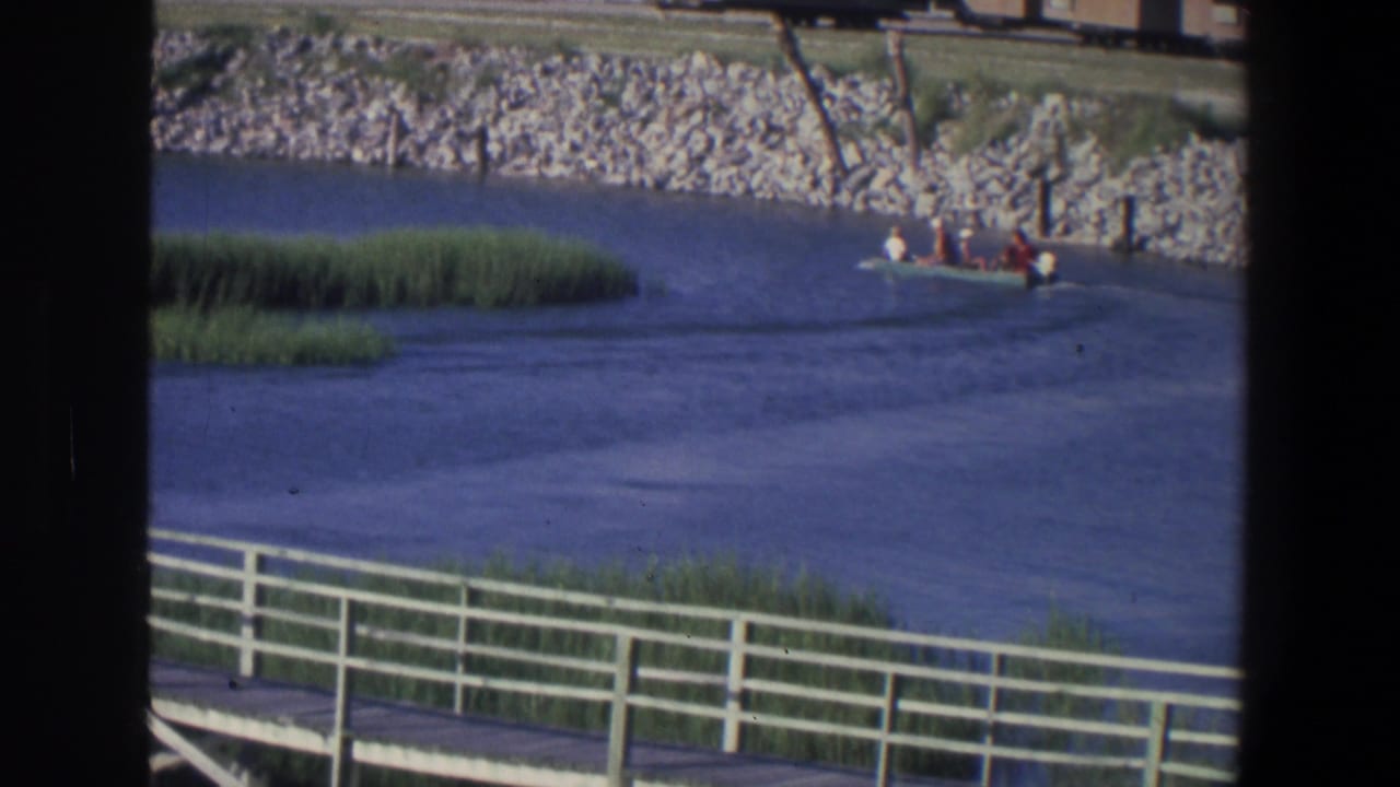 Small motorboat with passengers on calm river water near reeds and a railing.
