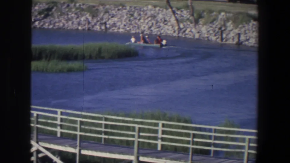 1971 Saint Paul home-movie frame with a distant motorboat and passengers beside reeds and a rocky embankment.