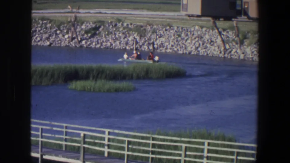 1971 Saint Paul 8mm frame of people in a small boat crossing the river scene near reeds and a riprap shoreline.