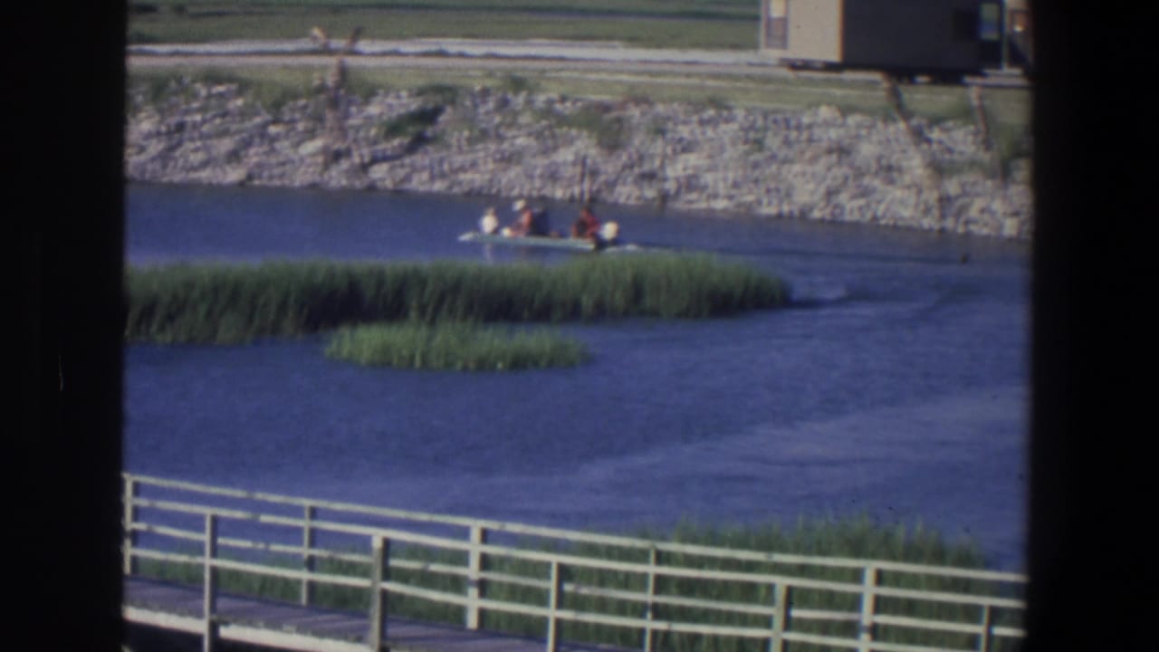 1971 Saint Paul home-movie frame showing a small motorboat moving past reeds on calm water, with a white pier railing and stone embankment.