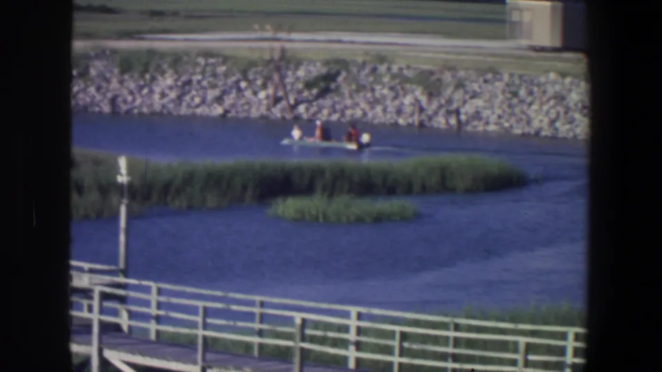 1971 Saint Paul frame with a small motorboat and distant passengers crossing blue water beside green reeds and a rocky bank.